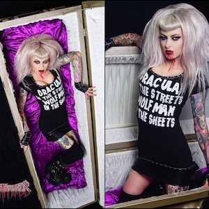 May include: A woman with pale skin and light hair, wearing a black dress with the text "DRACULA THE STREETS WOLF MAN IN THE SHEETS." She is in a coffin with purple velvet lining. The image has a gothic theme.