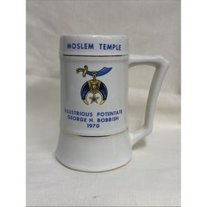 May include: A white ceramic beer stein featuring the blue text MOSLEM TEMPLE at the top and the text ILLUSTRIOUS POTENTATE GEORGE H. BOBBISH 1970 at the bottom. A central graphic depicts a blue scimitar above a crescent moon and a star.