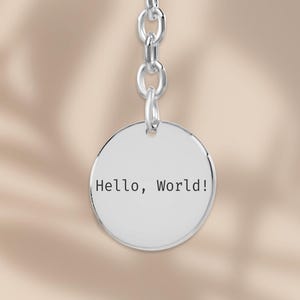 Könnte beinhalten: Ein silberner Schlüsselanhänger mit einem kreisförmigen Anhänger, der mit dem Text "Hello, World!" graviert ist. Der Schlüsselanhänger hat oben einen Ring und ist über eine kleine Kette mit dem Anhänger verbunden.
