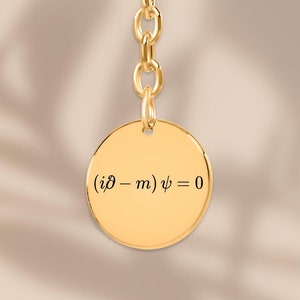 Op de afbeelding: Gouden sleutelhanger met een ronde hanger met de vergelijking '(i∂ - m)ψ = 0'.