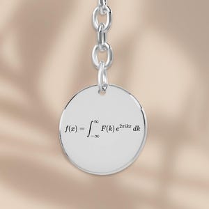Peut inclure: Un porte-clés en métal argenté composé d'un pendentif circulaire attaché à une courte chaîne et un anneau. Le pendentif est gravé avec la formule mathématique de la transformée de Fourier f(x) = intégrale de moins l'infini à l'infini F(k) e^2pikx dk.