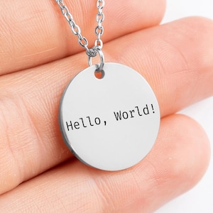 Peut inclure: Un collier argenté avec un pendentif rond portant l'inscription "Hello, World!". Le texte est en noir, en police sans-serif. La chaîne est fine et argentée.