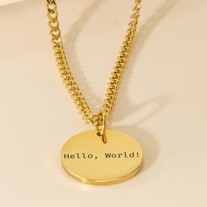May include: A gold-coloured necklace featuring a curb chain and a circular pendant. The pendant is inscribed with the text "Hello, World!". The necklace is presented against a plain, neutral backdrop.