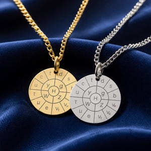 Pode incluir: Dois colares com pingentes de design circular. Um é dourado, o outro prateado. Cada pingente tem uma série de letras e símbolos dispostos em torno de uma letra central "H". Os colares estão pendurados em um tecido azul escuro.