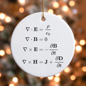 May include: A white, round Christmas ornament with a gold string for hanging. The ornament features black text with mathematical equations. The equations are related to electromagnetism. The background is blurred with warm, golden tones.