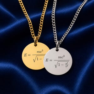 Op de afbeelding: Twee hangerkettingen met een natuurkundige formule op een donkerblauwe achtergrond. De ene ketting is goudkleurig, de andere zilverkleurig. Elke hanger is een ronde schijf met de formule E=mc²/√(1-v²/c²). De kettingen zijn van het curb-type.