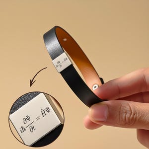 Op de afbeelding: Zwarte leren armband met een zilveren sluiting. De sluiting heeft een gegraveerde wiskundige vergelijking. De armband heeft een bruine binnenkant en een zilveren drukknoopsluiting.