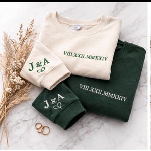 Puede incluir: Dos sudaderas y dos mangas, una crema y otra verde oscuro, bordadas con iniciales y una fecha en números romanos. Hay anillos dorados en la parte inferior. Las sudaderas están dobladas sobre una superficie de mármol.