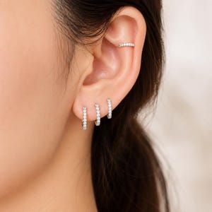 Puede incluir: Primer plano de una oreja adornada con varias joyas. Se ven tres pequeños pendientes de aro plateados, cada uno con pequeñas piedras transparentes. Un brazalete de oreja plateado a juego, también engastado con piedras transparentes, se lleva más arriba en la oreja.