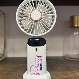 May include: A white and rose gold portable fan with the text "Pretty Moody" in a script font. The fan has a circular head with white blades and a rose gold accent. The base is white and has a unique shape.