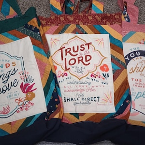 May include: Three fabric tote bags featuring quilted patchwork designs in various colors. Each bag has a central white panel with printed religious text and floral illustrations. The text includes phrases like Set Your Minds On Things Above, Trust in the Lord, and The Lord You & Keep You.
