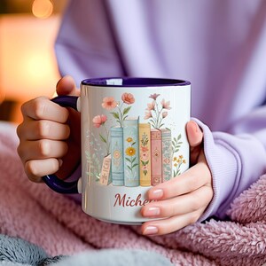 May include: White ceramic mug with a purple interior and handle, featuring a floral and book design. The design includes pastel-colored books with flowers and the name "Miche" in a script font. The mug is held by a person.