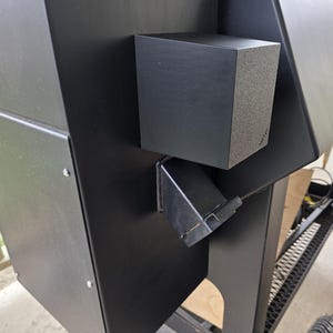 May include: Black outdoor smoker with a square side box and a metal chute. The smoker has a matte finish and is designed for grilling and smoking food. The smoker is on wheels.