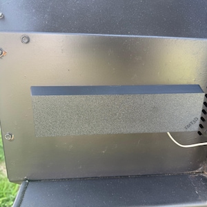 May include: Close-up of a black metal object with a textured, rectangular panel. The panel has a dark gray surface and the text "TMT3D" is visible. A white wire extends from the object.