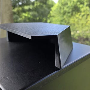 May include: A black metal mailbox with a textured finish. The mailbox has a sloped top and a rectangular body. The background shows green foliage and a window.