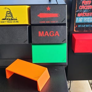 May include: A black smoker with various colored accents. The smoker has a yellow box with a snake graphic and the text "DON'T TREAD ON ME". Other boxes include the text "COME AND TAKE IT" and "MAGA". An orange shelf is in the foreground.