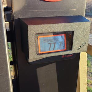 May include: A black rectangular device with a digital display showing the number 77. The display has an orange border and a blue background. The device is mounted on a larger black surface. The text "FireBoard" is visible.