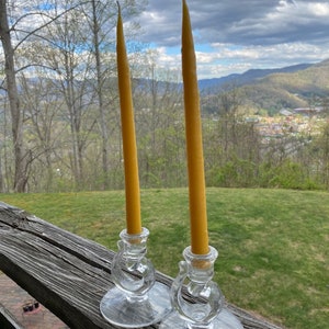 May include: Two tall, yellow beeswax candles in clear glass candle holders with a decorative design. The candles are on a wooden surface with a green grassy background.