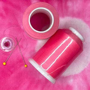 May include: A large spool of vibrant pink sewing thread, a smaller clear plastic bobbin with matching pink thread, and two long sewing pins with yellow spherical heads. The items are arranged together, showcasing the bright pink color and texture.
