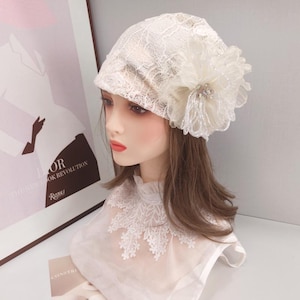 May include: A cream-coloured lace beanie hat featuring a large, layered fabric flower embellishment on the side. The flower is accented with clear beads and sequins. The hat is displayed on a mannequin head wearing a white lace-trimmed collar.