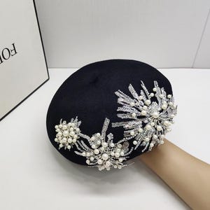 Puede incluir: Un sombrero boina negro con elaborados adornos florales. Las decoraciones presentan racimos de perlas de imitación y brillantes pedrería plateada. El sombrero es un accesorio elegante para ocasiones especiales.