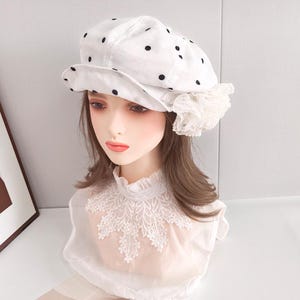 May include: A white newsboy cap featuring a black polka dot pattern and a ruffled white lace accent on the side. The cap has a structured brim and is displayed on a mannequin head wearing a white collar with intricate floral lace embroidery.