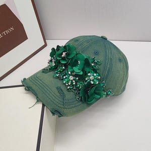 May include: A green denim baseball cap with a large green floral embellishment. The flowers are made of fabric and have rhinestones in the center.