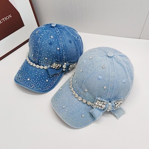 Puede incluir: Dos gorras de béisbol de mezclilla, una azul oscuro y otra azul claro. Ambas están adornadas con pedrería y detalles en forma de estrella. Un lazo decorativo y una hilera de pedrería adornan la visera de cada gorra.