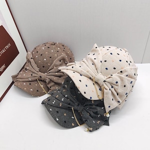 Peut inclure: Trois casquettes de baseball en beige, marron et gris foncé, chacune dotée d'un grand nœud en tissu sur le devant, d'une fine chaîne dorée drapée sur le nœud et d'une petite perle blanche. Les casquettes présentent un motif à pois texturé.