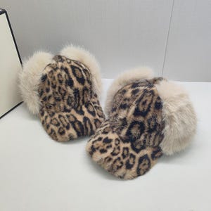 May include: Two leopard print baseball caps with cream-coloured fur trim. The caps feature a brown and black spotted pattern. The fur trim is around the ear area of the caps. The caps are on a white surface.