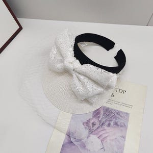 May include: A white headband with a large sequined bow and a delicate veil. The headband has a black band and a white, woven base. The bow is covered in small, shiny sequins. The veil is a fine, sheer fabric.