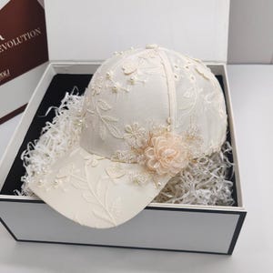 May include: Cream-colored baseball cap with intricate floral embroidery and pearl accents. The cap features a curved brim and is adorned with a delicate peach-colored flower and sparkling embellishments. Presented in a white gift box with black trim.