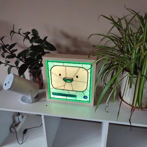 May include: A stained glass lamp shaped like a computer screen with a cartoon face. The lamp is lit up with a green glow and is sitting on a white shelf.