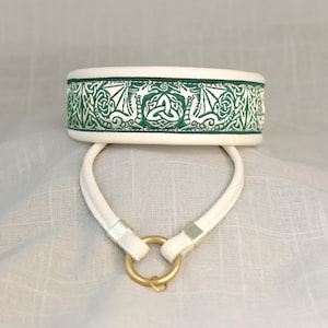 Könnte beinhalten: Ein weißes Halsband mit einem grünen keltischen Knotenmuster, das auf das Band gestickt ist. Das Halsband hat einen weißen Riemen mit einem goldfarbenen Ring. Das Design ist ein sich wiederholendes Muster aus verschlungenen Linien und Formen.
