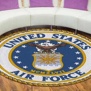 Op de afbeelding: Een ronde vloerkleed met het embleem van de US Air Force. Het embleem staat centraal op een blauwe achtergrond met witte sterren en een gouden rand. Het kleed ligt op een houten vloer voor een witte bank met paarse kussens.