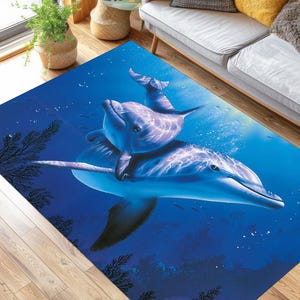 Pode incluir: Um tapete azul e cinza com um desenho de dois golfinhos nadando no oceano. Os golfinhos estão em uma pose brincalhona, com um golfinho pulando sobre o outro. O tapete tem um fundo azul com bolhas brancas e alguns peixes pequenos.