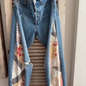 Może przedstawiać: Jasnoniebieskie dżinsy z panelami z patchworku po bokach. Patchwork ma kwiatowe i geometryczne wzory w odcieniach czerwieni, żółci i kremu. Dżinsy mają zapięcie na guziki i postrzępione obszycia.