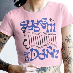 Puede incluir: Top corto rosa con el texto "SYSTEM OF A DOWN" en azul. La camiseta tiene un diseño gráfico con esposas, un código de barras y una cerradura. La modelo lleva una falda a rayas blancas y negras.