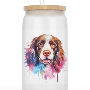 Könnte beinhalten: Ein weißer Glasbecher mit Holzdeckel und einem durchsichtigen Strohhalm. Der Becher zeigt ein Porträt eines Hundes im Aquarellstil mit braunem und weißem Fell sowie farbenfrohen Spritzern in Rosa, Blau und Lila.
