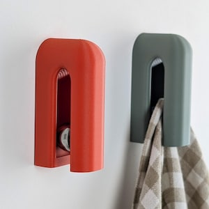 Può includere: Tre ganci da parete in grigio, rosso e verde. I ganci sono progettati con una forma ad arco arrotondata e una fessura al centro. Un asciugamano da cucina è appeso al gancio verde.