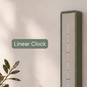 May include: A vertical linear clock with a sage green frame and a central display panel. The display features a series of small, illuminated dots in white, pink, and blue, arranged in a column with black horizontal markers. The text Linear Clock is visible.