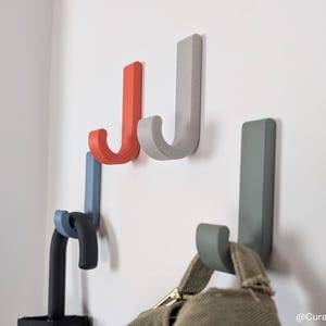 Puede incluir: Ganchos de pared con forma de "J" en varios colores, incluyendo rojo, gris, azul y verde. Los ganchos están montados en una pared blanca. Un sombrero cuelga de uno de los ganchos. Los ganchos son de material sólido.