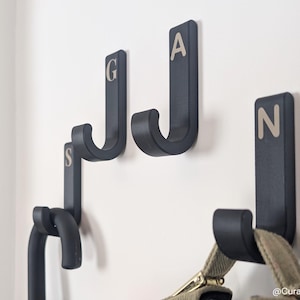 Puede incluir: Cuatro perchas de pared de color gris oscuro con las letras G, A, S y N en color claro. Los ganchos están montados en una pared blanca, con una bolsa colgando del gancho con la letra N.