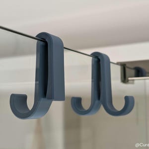 Puede incluir: Tres ganchos de plástico azul están montados en una puerta de ducha de vidrio. Los ganchos tienen forma de letra J y tienen un extremo redondeado para colgar objetos.