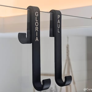 Peut inclure: Deux crochets noirs en forme de J suspendus à une barre métallique. Le crochet de gauche porte le nom "GLORIA" et celui de droite "PAUL". Ces crochets sont conçus pour suspendre des objets.