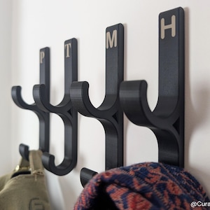 Puede incluir: Un conjunto de cuatro ganchos de pared negros, cada uno con un vástago vertical con un gancho curvo y una letra en la parte superior. Las letras P, T, M y H están impresas en un color claro en la parte superior de cada gancho.