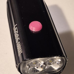 Puede incluir: Luz de bicicleta Lezyne Lite Drive 1000XL rectangular negra con un botón rosa. La luz tiene dos bombillas LED brillantes y está diseñada para ciclismo. El nombre del producto está impreso en blanco.