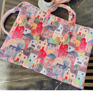 Op de afbeelding: Een roze laptoptas met een kleurrijke print van huizen en bomen. De print is in aquarelstijl met een speels gevoel.
