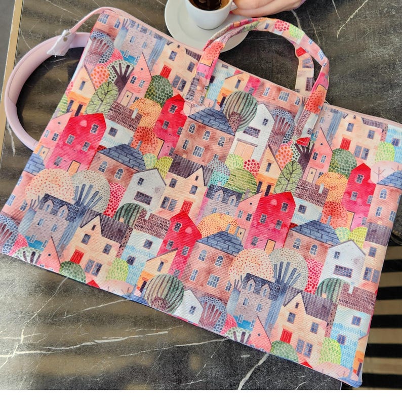 Op de afbeelding: Een roze laptoptas met een kleurrijke print van huizen en bomen. De print is in aquarelstijl met een speels gevoel.