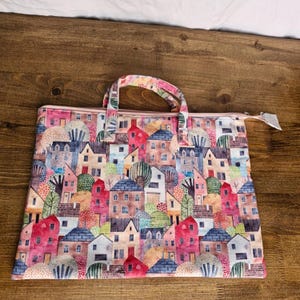 Op de afbeelding: Een roze tote bag met rits met een aquarelprint van kleurrijke huizen en bomen. De tas heeft twee handvatten.
